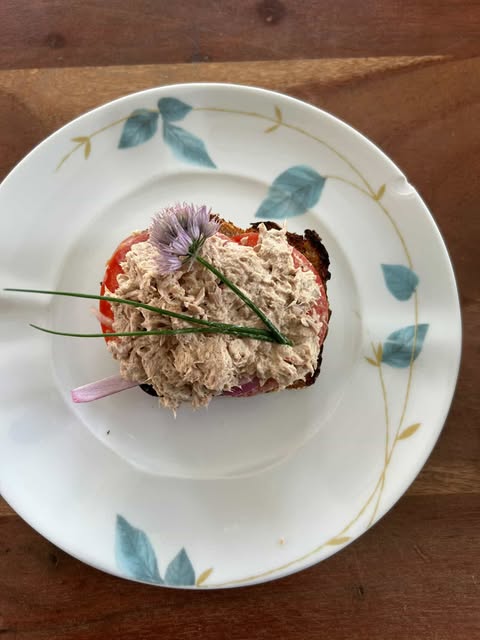 Open-faced tuna salad sandwich on toasted bread, topped with a slice of tomato, garnished with fresh chives and a chive blossom, served on a white plate with a blue and gold leaf pattern.