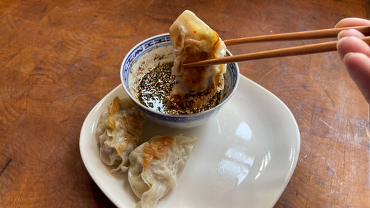 a dumpling being held in chopsticks over a bowl of dipping sauce on a white plate with 2 other dumplings