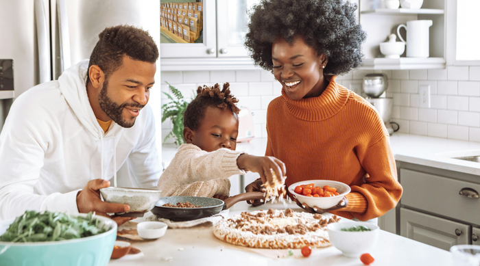 a family of 3 in a bright kitchen, sitting at the kitchen table topping a homemade pizza, with the young child sprinkling on the cheese as the mother and father hold bowls of toppings.