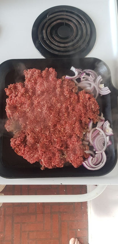 Large square cast iron griddle filled with raw ground beef and in 2 corners sliced red onions just starting to cook  on a white electric stove top with one element visible 