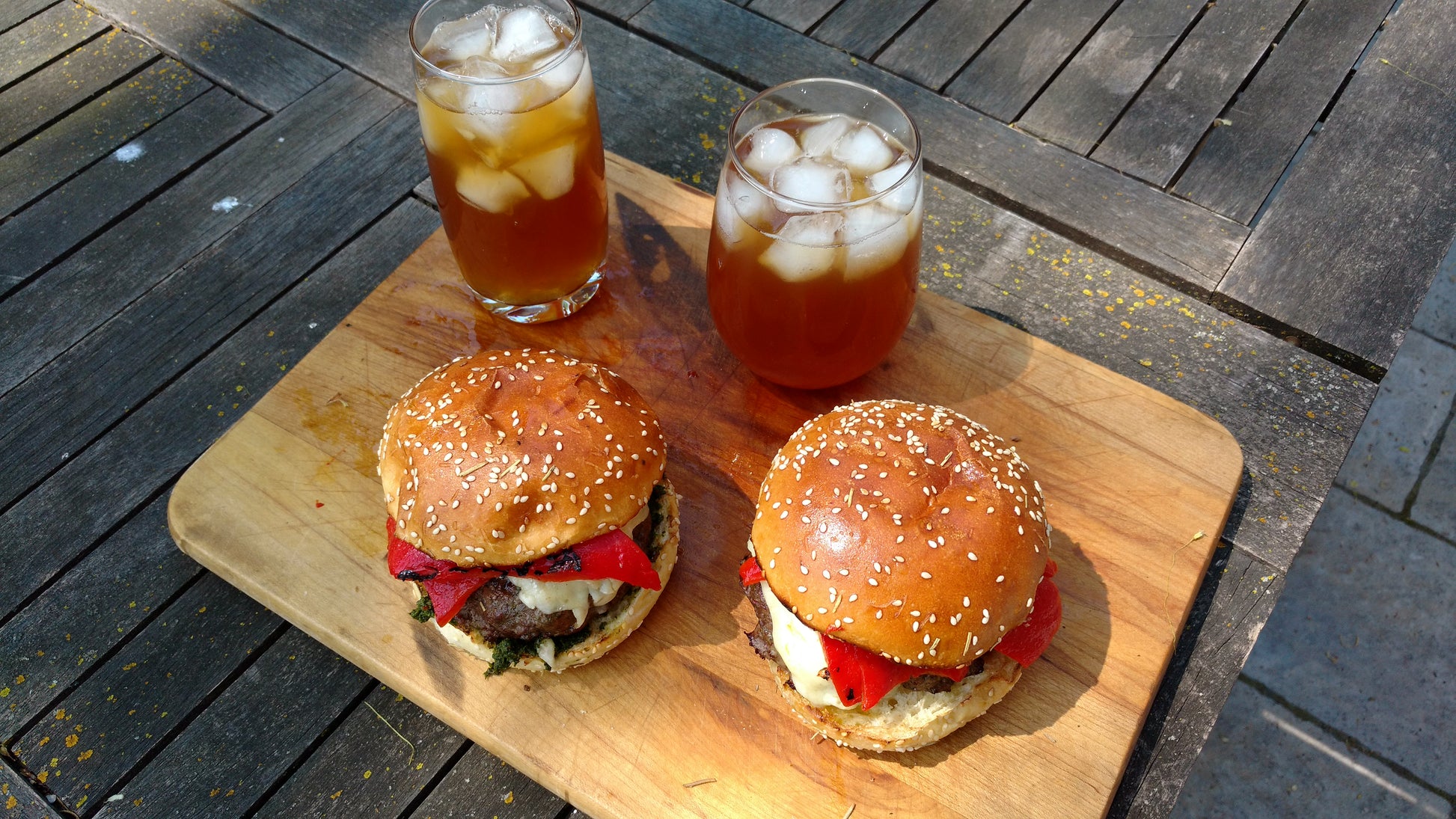 2 burgers with sesame seed buns served on a wooden cutting board with 2 glasses filled with ice and iced tea 